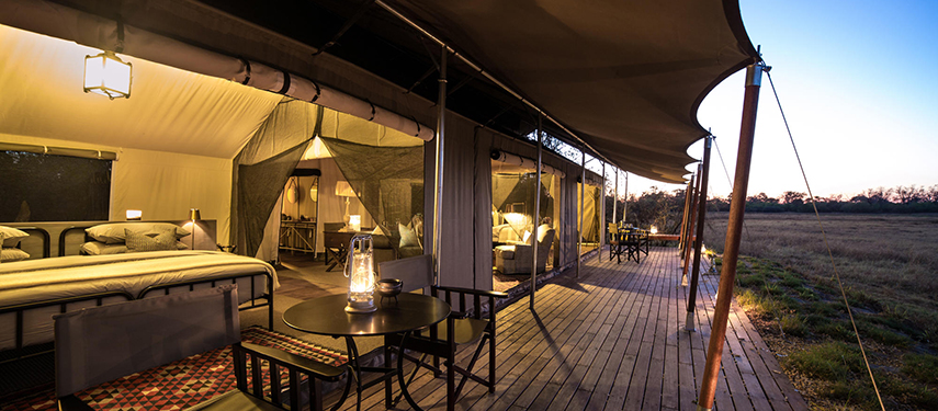 Deck view of a tented suite at dusk, featuring canvas flaps, seating, and soft interior lighting spilling onto the wooden platform.
