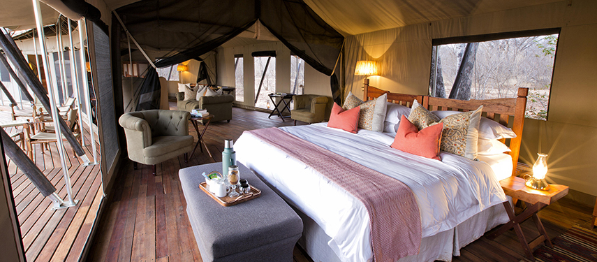 Charming tented suite with a central bed, elegant wooden furniture and natural light flooding in through large windows.
