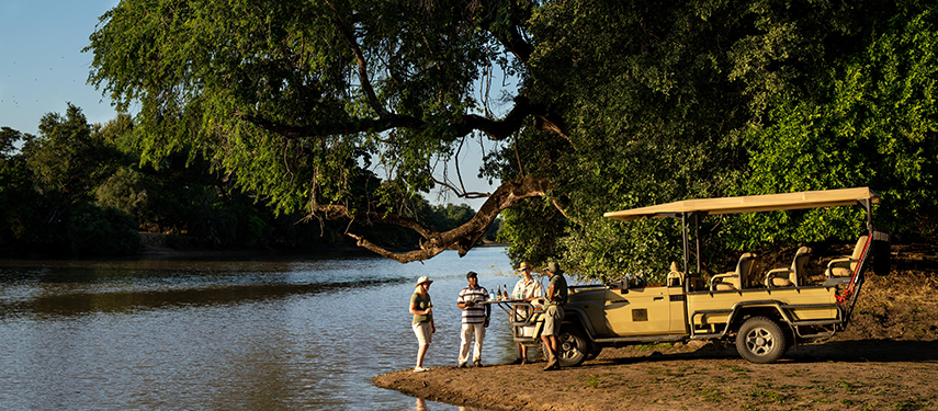 A group gathers for sunset drinks beside the river, with a safari vehicle parked under a sprawling tree canopy.