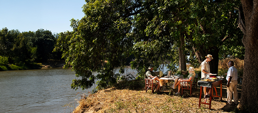 Guests dine under shady trees on the banks of the Zambezi, enjoying a relaxed bush lunch prepared al fresco.