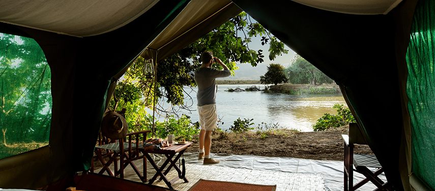 A guest stands at the edge of their tent, gazing across the river with binoculars as hippos rest in the shallows.