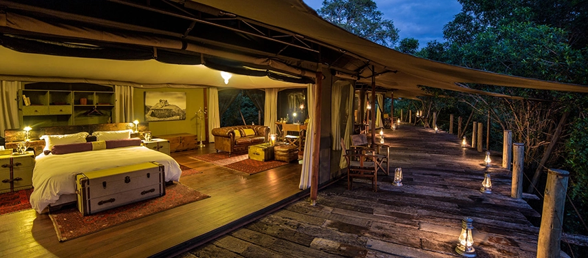 Interior of a Mara Plains safari suite in Kenya, featuring an open-plan bedroom, elegant furnishings, and lantern-lit decking overlooking the wilderness.