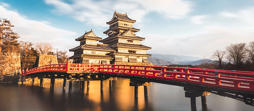 Matsumoto Castle viewed across a red bridge at sunset, highlighting its dramatic black architecture and mountain backdrop.