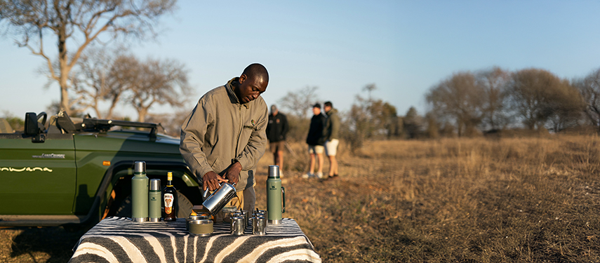 Ranger prepares morning coffee and Amarula on a table set beside a safari vehicle during a game drive stop.