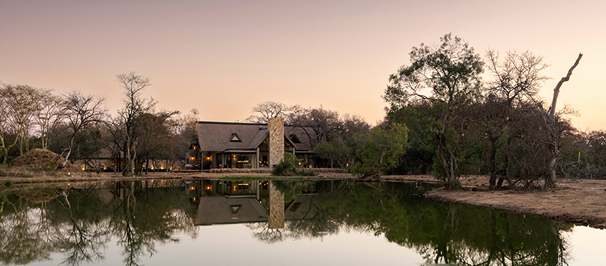 Serene view of Monwana Lodge reflected in a nearby waterhole at dawn, with soft pastel skies overhead.