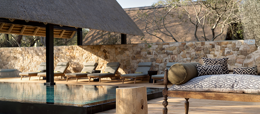 Stylish poolside area with cushioned loungers set against a stone wall and thatched pavilion.