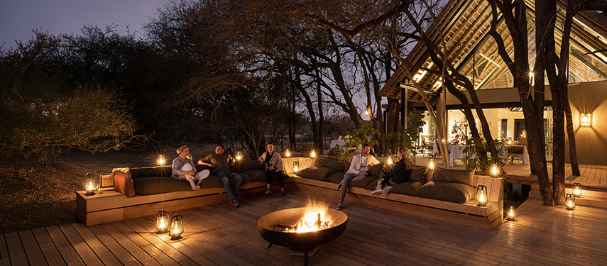 Inviting indoor lounge area with a roaring stone fireplace and expansive bushveld views through large windows.