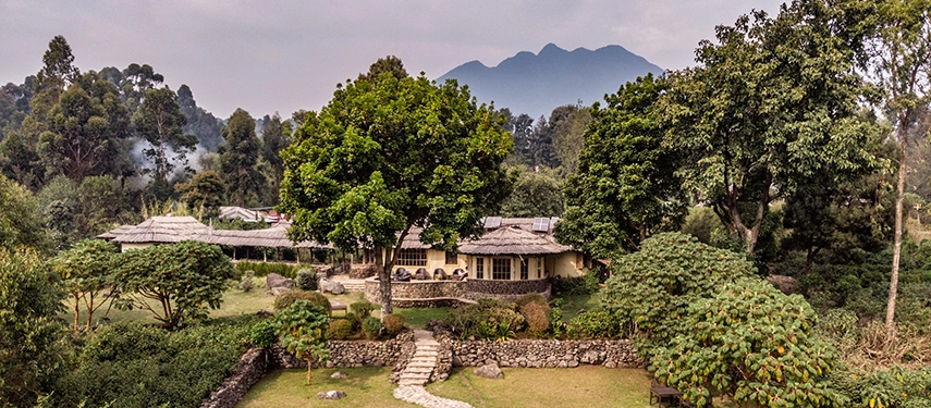 Aerial view of Mount Gahinga Lodge surrounded by lush gardens and forest with the Virunga volcanoes in the background.