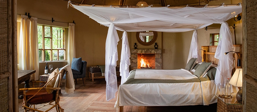 Elegant guest banda interior featuring a four-poster bed with mosquito netting, stone fireplace, and rustic furnishings.