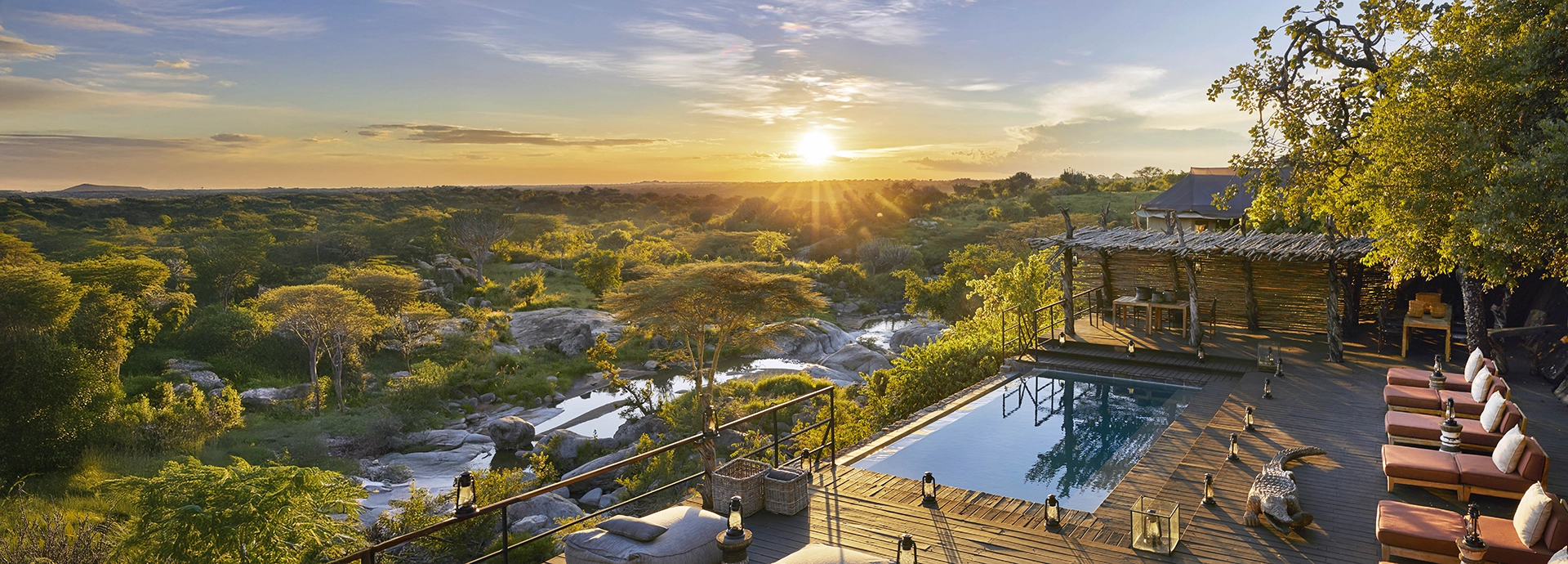 Sunset view over the main pool area at Mwiba Lodge in the Serengeti, with a timber deck, lantern-lit loungers and sweeping views across the rocky wilderness and river below.