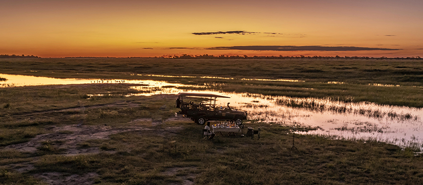 Sundowners set up beside a safari vehicle amidst golden floodplain reflections during sunset in the Chobe wilderness.