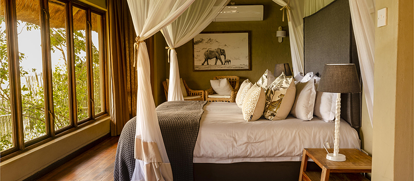 A stylish bedroom at Ngoma Safari Lodge with a canopy bed and earthy decor overlooking the surrounding trees.