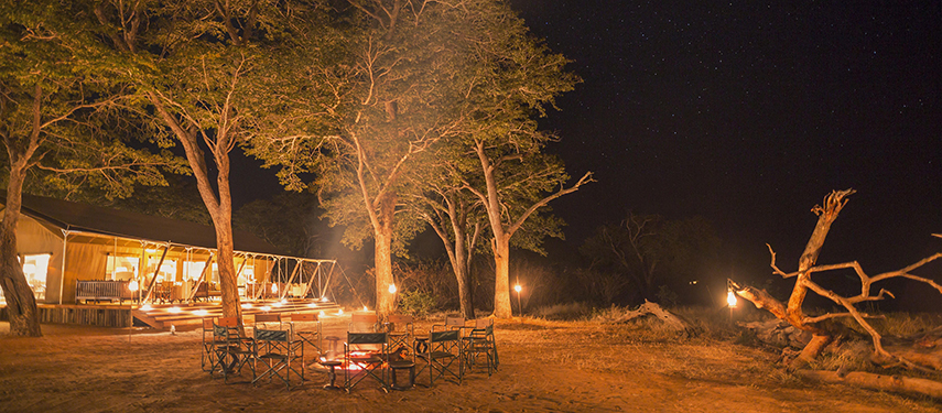 Campfire glows warmly under the stars with chairs set in a circle and the softly lit tented lodge in the background.