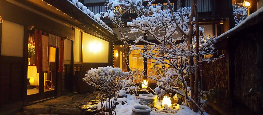 A lantern-lit courtyard at a traditional ryokan in winter, where fresh snow enhances the warmth and intimacy of the setting.