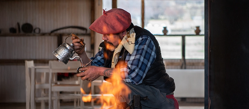 A gaucho kneels by a fire, carefully pouring hot water for maté inside a rustic Patagonian quincho.