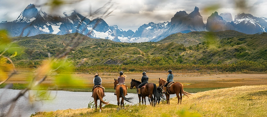 Riders pause beside a still Patagonian lagoon, gazing towards the dramatic granite towers and snow-lined peaks of Torres del Paine.