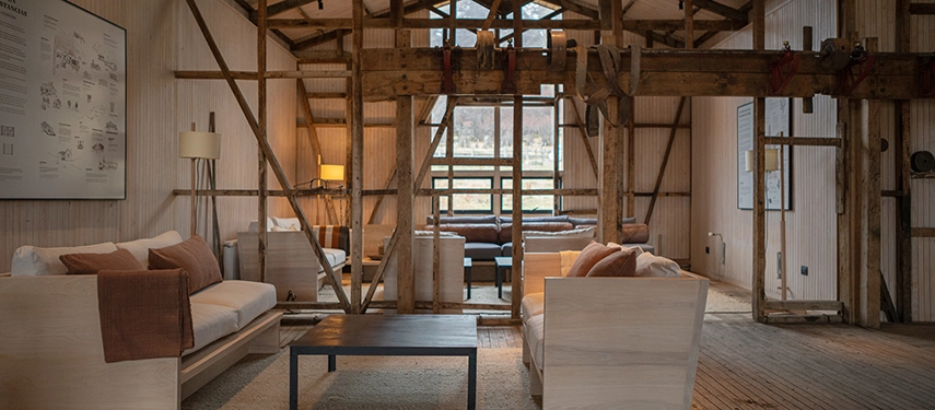 A rustic lodge interior features exposed beams and warm timber textures, with soft seating arranged around a central lounge space.