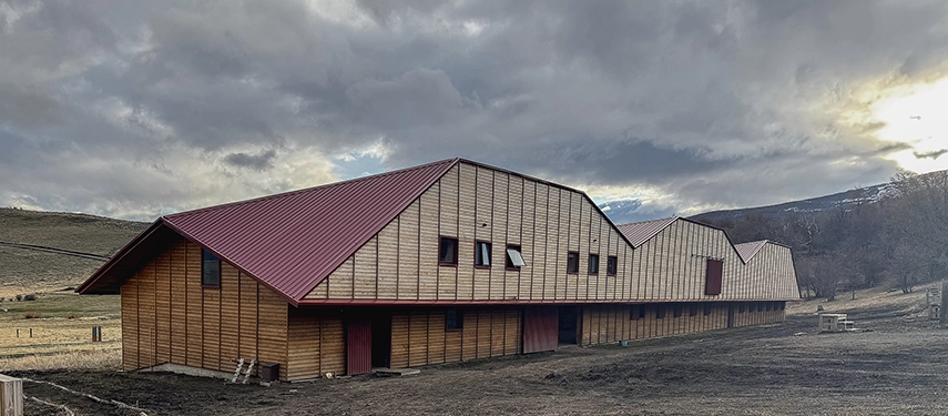A long timber stable building stands beneath brooding Patagonian skies, its red roof contrasting with the rugged landscape.