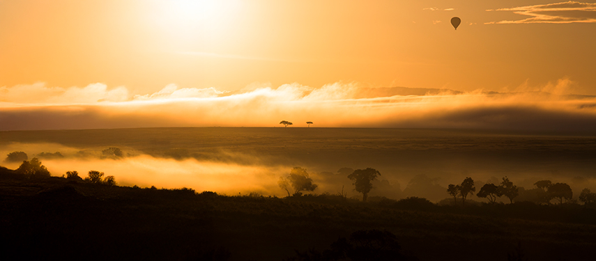 Golden sunrise over the Masai Mara with a hot air balloon floating above mist-covered plains.