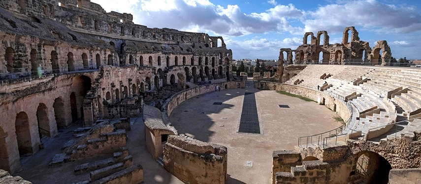 The Roman amphitheatre of El Jem with towering stone arches and tiered seating under a cloudy sky.