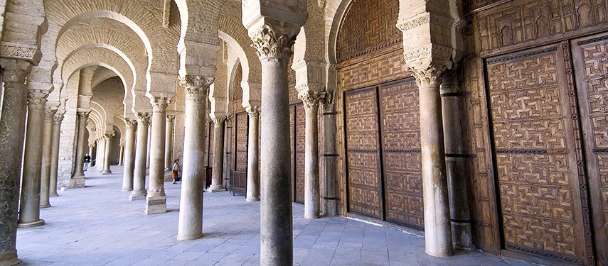 Exterior arcade of Kairouan’s Great Mosque with arched stone columns and intricately carved wooden doors.
