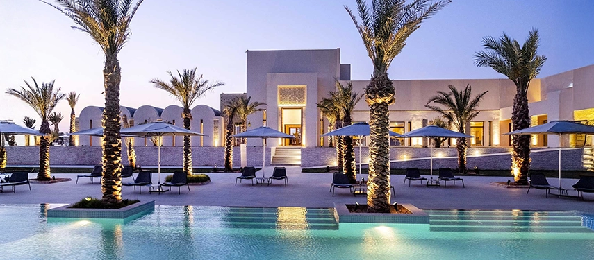 Evening view of The Residence Douz pool area illuminated by soft lighting and surrounded by palm trees.