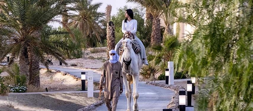A woman riding a camel through the palm-lined pathways of The Residence Douz gardens.
