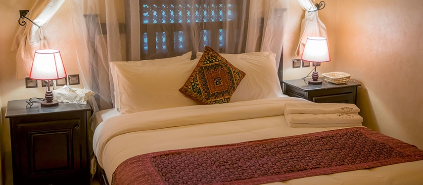 Intimate Moroccan bedroom with soft lighting, wooden furniture, and embroidered fabrics on the bed.