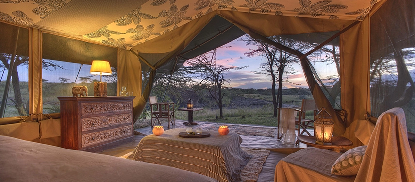 A luxury canvas tent bedroom at Hemingways River Camp Mara, glowing with warm lamplight and candles as the sun sets over the Kenyan bush beyond.