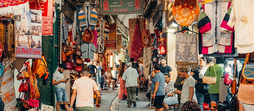 Bustling souk in Tunis Medina with colourful textiles, leather goods, and shoppers beneath vaulted brick arches.