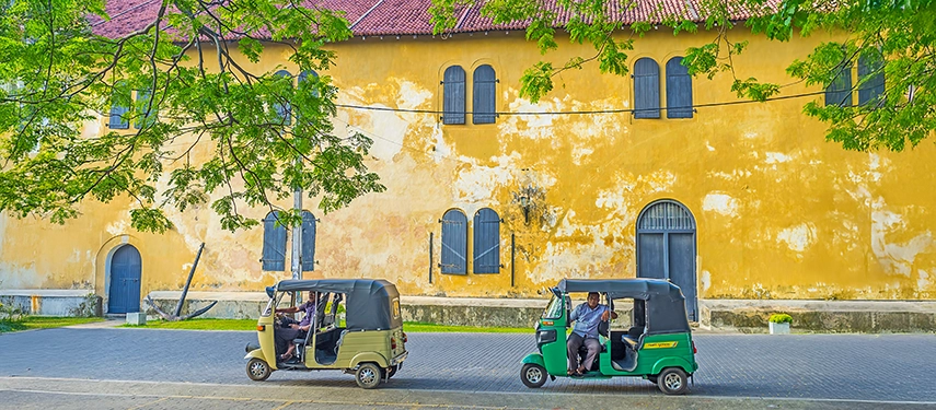 Colourful tuk-tuks parked along a street in front of a rustic yellow colonial building in Sri Lanka.