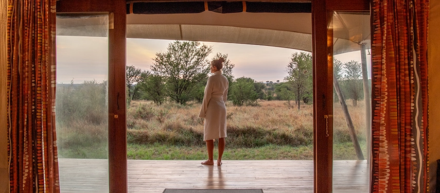 Guest standing on a private deck in robe enjoying sunrise views from Sayari Camp tent in the Serengeti.
