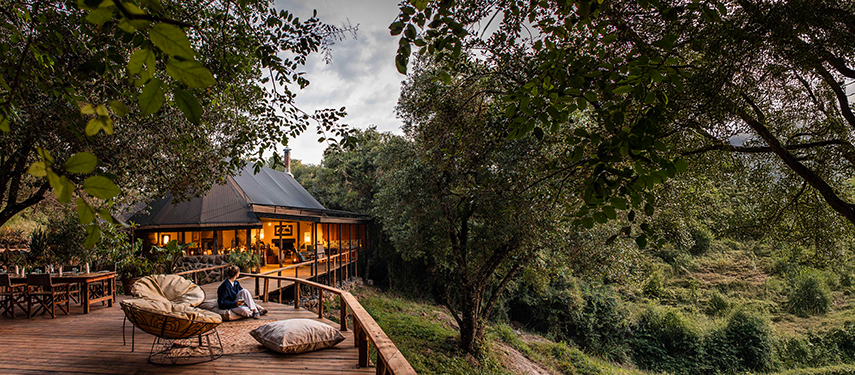 Woman sitting on a cushioned chair overlooking the forest, with the warmly lit main mess tent of Serian ‘The Original’ in the background.