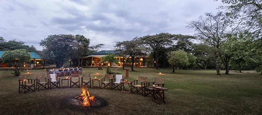 Camp chairs arranged in a semicircle around a crackling fire at Hemingways River Camp Mara at dusk, with a long dining table set beneath the trees and the illuminated mess tent beyond.