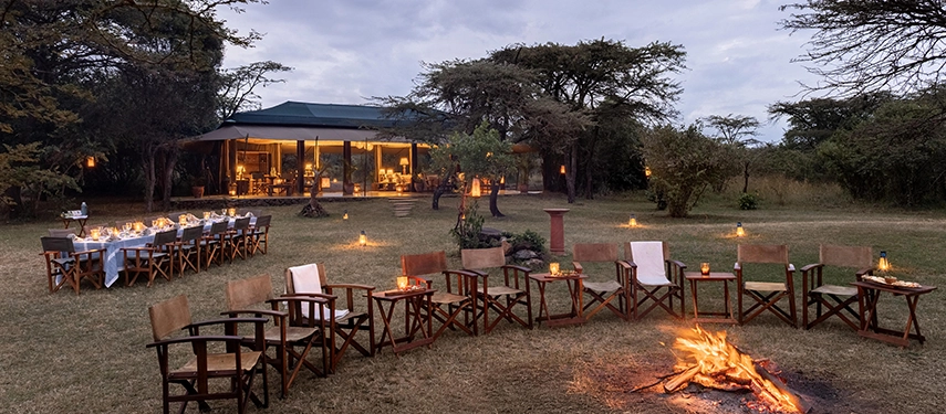 A candlelit outdoor dining table and circle of camp chairs arranged around a crackling fire at Hemingways River Camp Mara, with the illuminated mess tent glowing softly in the background.