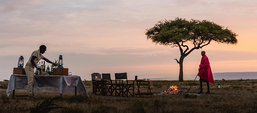 A Maasai warrior in a red shuka tends a fire on the open plains at sunset while a camp attendant prepares sundowner drinks beneath a solitary acacia tree.