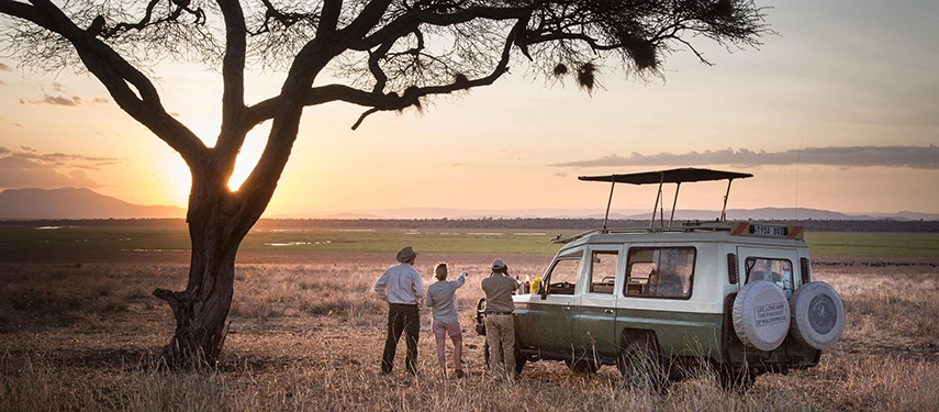 Guests gather beside a safari vehicle to enjoy sundowners as the sun sets across vast plains and distant escarpments.