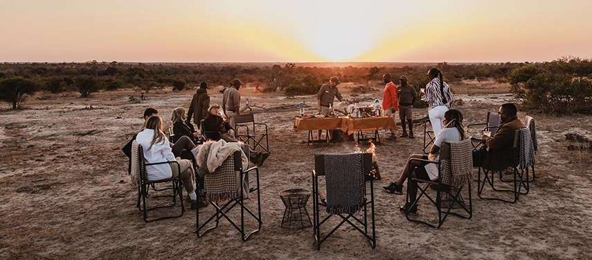 Guests sitting in a circle around a fire in a dry riverbed enjoying a bush dinner at sunset.