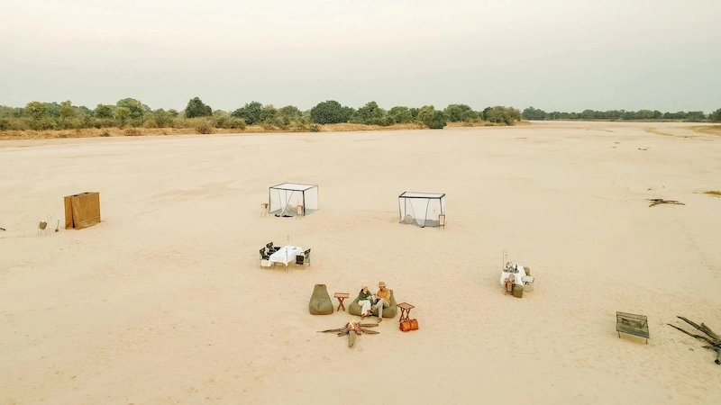 Aerial view of an intimate bush dinner at Time + Tide Luwi Camp in South Luangwa National Park, set on a wide sandy riverbed with lantern-lit tables.