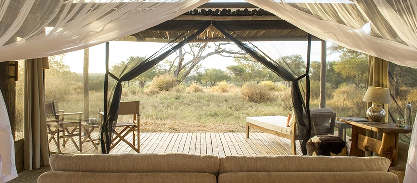 A shaded canvas suite opens onto a wide wooden deck with uninterrupted views across golden grasslands and scattered acacia trees.