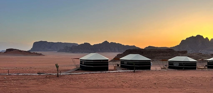 Traditional Bedouin tents stand quietly in the open desert as the sky fades from gold to blue behind distant mountains.
