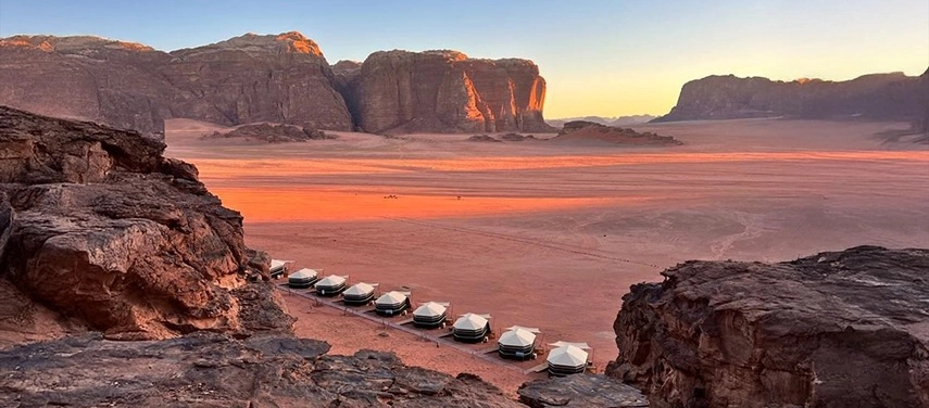 An elevated view reveals a line of desert tents stretching across the red sands, framed by towering sandstone cliffs at golden hour.