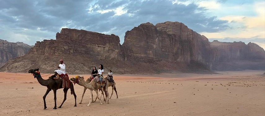 Guests ride camels across the open sands of Wadi Rum, guided by Bedouin hosts against a backdrop of monumental cliffs.