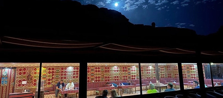 The open sided dining tent glows warmly at night, with traditional Bedouin textiles and guests gathered beneath a moonlit desert sky.