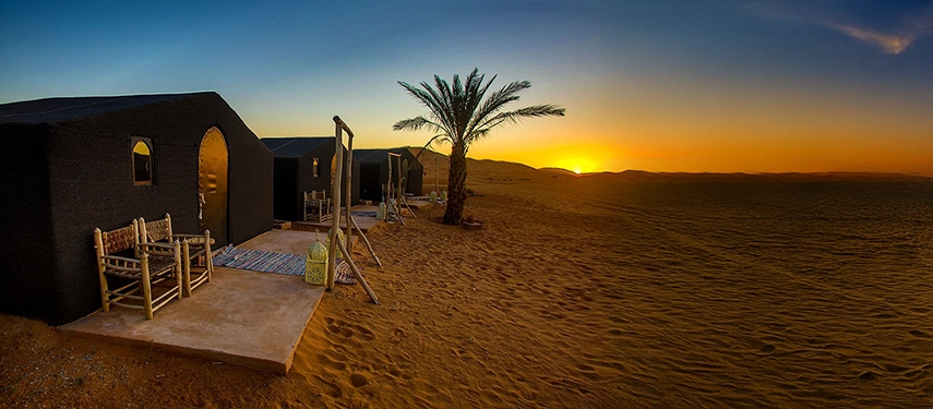 Row of luxury tents with palm trees at sunset in the Sahara desert at Riad Madu camp.