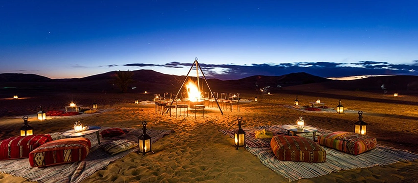 Magical outdoor campfire setup with lanterns, cushions, and rugs beneath a twilight desert sky.