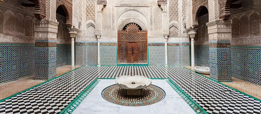 Traditional Moroccan courtyard at Al Attarine Madrasa with ornate tilework, carved stone arches, and a central fountain.