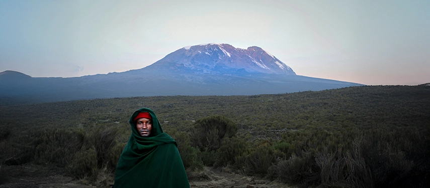 Local guide wrapped in a green shuka standing before the vast foothills of Mount Kilimanjaro at dusk.