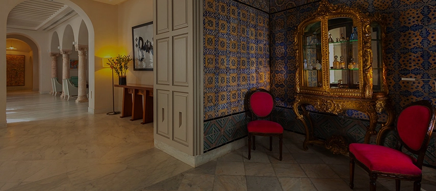 Elegant hotel corridor with Tunisian tilework, ornate gold display cabinet, and red velvet chairs.