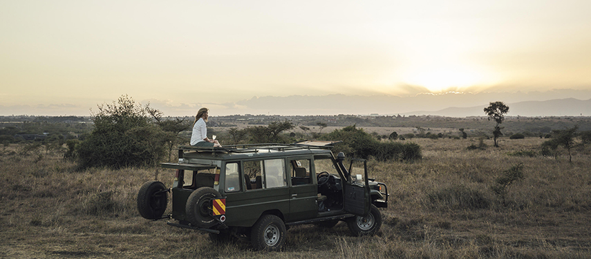 A woman sits on top of a safari vehicle at sunrise, overlooking the plains of Nairobi National Park.